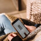High angle of unrecognizable woman sitting on chair and choosing clothes in internet shop using mobile phone